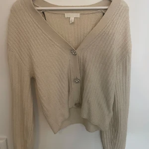 Beige ribbad kofta med blommiga knappar - Superfin beige ribbad kofta från H&M med v-ringning och långa ärmar. Snygga blommiga knappar med glittrande stenar ger en extra touch. Perfekt att slänga över en topp för en chill men ändå stylish look. Går att diskutera pris 