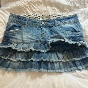 Volangig jeanskjol från DN Jeans - Supersöt blå jeanskjol med flera volanger och fransiga kanter. Klassisk femficksmodell med bälteshällor och detaljer i silver. Perfekt för dig som vill ha en cool och lekfull stil. Innerlinning med textprint. Snygg till sneakers eller boots!