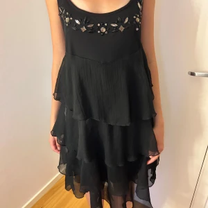 Svart volangklänning med pärldetaljer - Supersnygg svart klänning med flera lager volanger och pärl- och stenbroderier vid urringningen. 
