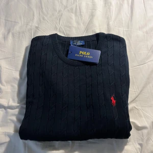 Svart kabelstickad tröja Polo Ralph Lauren - Kabelstickad svart tröja från Polo Ralph Lauren med klassisk rund halsringning och röd broderad logga på bröstet. Långärmad modell i mjuk bomull, perfekt för dig som gillar stilren och tidlös design.