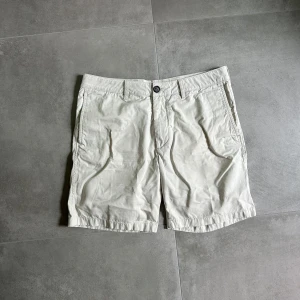 Beige bomullsshorts från Arket - Snygga beige shorts från Arket i ekologisk bomull. Klassisk modell med fickor fram och bak, bälteshällor och knappstängning. Perfekta för varma dagar och enkel att matcha med allt. Stilren och clean look. Artnr: B355