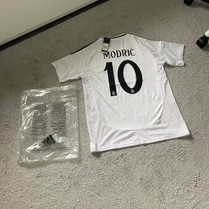 Real Madrid Modric 10 Adidas tröja - Säljer en officiell Real Madrid fotbollströja från Adidas med Modric 10 på ryggen. Tröjan är vit med svarta detaljer, klubbmärke och sponsortryck på bröstet. Tillverkad i lätt och ventilerande material, perfekt för match eller träning.