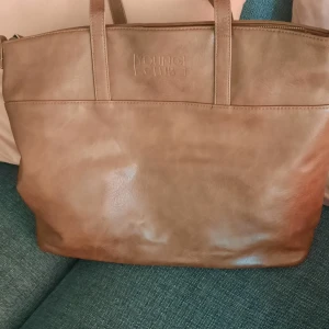 Brun axelväska från Young Club - Snygg brun axelväska från Young Club i slätt skinn. Väskan har två rejäla handtag och enkel, stilren design utan mönster. Perfekt storlek för att få plats med allt du behöver till skolan eller stan.