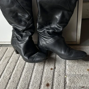 Svarta stövlar med klack, storlek 38 - Snygga svarta stövlar i skinn med rund tå och stabil klack. De har en klassisk design med lätt rynkade skaft och passar perfekt till både jeans och kjol. Skorna är helsvarta och har en tidlös look som funkar året runt.