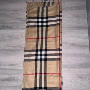 Beige rutig halsduk från Burberry - Klassisk halsduk från Burberry i beige med det ikoniska rutmönstret i svart, vitt och rött. Tunn och mjuk, perfekt att slänga över axlarna för en snygg touch. Fransar i kanterna och diskret Burberry-märke. En riktig stilhöjare!