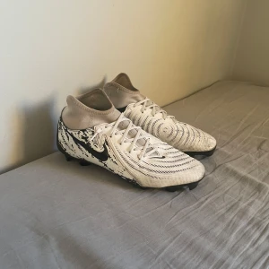 Nike vita fotbollsskor med svart mönster - Säljer ett par vita egen designade Nike fotbollsskor med svart grafiskt mönster på hälen och sidorna. Skorna har snörning, en strumpliknande krage. Perfekta för dig som vill sticka ut på planen med stilren design och bra grepp. Ny pris 1700 kr. hör av er om ni har frågor. 