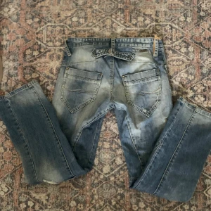 Blå slitna jeans med raka ben - Säljer ett par blå jeans med coola slitningar och tvättade detaljer. Jeansen har raka ben och snygga bakfickor, perfekta för en avslappnad niche stil. Köpta secondhand men aldrig använda av mig och bra skick med riktig denim!