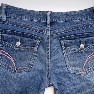 Lågmidjade y2k jeans - Snygga blå vintage jeans från låg midja. Jeansen har coola detaljer som rosa broderi och ett litet hjärta på myntfickan. Jeansen är knappt använda och är i storlek W29, längd 30