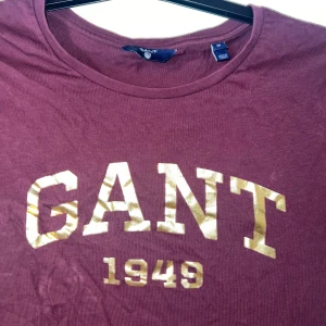 Vinröd GANT t-shirt med guldtryck - Vinröd t-shirt från GANT med korta ärmar och rund hals. Framsidan har ett stort, glansigt guldtryck med texten 'GANT 1949'. Skön och klassisk passform i mjuk bomull, perfekt för en chill och stilren look. Passar xs-L❤️