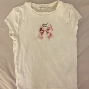 Vit ring topp med rosett från H&M - Supersöt vit ribbad topp från H&M med korta ärmar. Framtill finns ett tryck med en rosa rosett och texten 'Kind Heart' ovanför. Perfekt för dig som gillar gulliga detaljer och enkel stil.