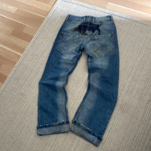 Rusty Neal blå jeans med broderad tjur - Säljer ett par blå jeans från Rusty Neal, modell South Dakota. Jeansen har en broderad svart tjur på bakfickan, slitna detaljer och uppvikta benslut. Klassisk femficksmodell med orange sömmar och cool vintagekänsla. Perfekta för dig som gillar streetwear.
