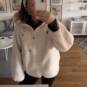 Beige teddyjacka med dragkedja - Supermysig beige teddyjacka med huva och dragkedja framtill. Jackan har en bröstficka med dragkedja och oversized passform som ger en chill vibe. Perfekt för kalla dagar när du vill vara både varm och snygg.