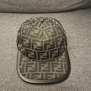 Fendi keps med FF-mönster - Snygg keps från Fendi med ikoniskt FF-logomönster i svart och grått. Kepsen har böjd skärm med svart kant och justerbar rem bak. Tillverkad i ett slitstarkt tyg som känns lyxigt och trendigt. Perfekt accessoar för att lyfta din streetwear-look.