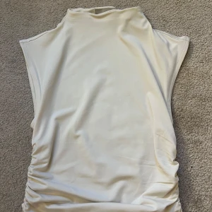 Vit ärmlös topp från H&M XS - Säljer en stilren vit ärmlös topp från H&M i storlek XS. Toppen har hög hals och lätt rynkade sidor för en snygg passform. Perfekt att matcha med jeans eller kjol för en clean look. Aldrig använd.