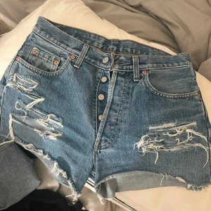 Levi's slitna jeansshorts hög midja - Snygga blå jeansshorts från Levi's med hög midja och slitna detaljer framtill. Klassisk femficksmodell, knappar i gylfen och råa kanter nertill för en avslappnad vibe. Perfekta för sommaren och ger en cool, vintage känsla.