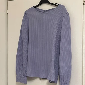 Ljuslila randig blus med långa ärmar - Supersöt ljuslila blus med vertikala ränder och långa puffiga ärmar. Blusen har breda muddar vid ärmsluten och en rund halsringning. Perfekt för dig som gillar en stilren men ändå trendig look.