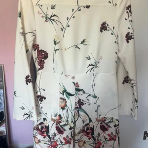 Vit blommig långärmad klänning - Superfin vit klänning med blommigt mönster i rött, grönt och beige. Långärmad modell med rak passform och diskret söm i midjan. Perfekt för vår och sommar när du vill känna dig extra fräsch och stilren.