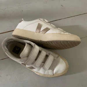 Vita Veja sneakers med guldiga detaljer - Snygga vita sneakers från Veja med guldiga detaljer och klassisk V-logga på sidan. Skorna har tre kardborreband för enkel påtagning och en platt sula med hexagonmönster undertill. Tillverkade i skinn och passar perfekt till en clean och stilren look.
