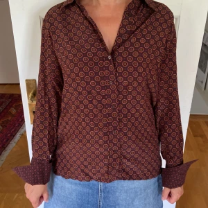 Brun mönstrad skjorta med knappar - Snygg brun skjorta med cirkelmönster i orange och gult. Skjortan har klassisk krage, långa ärmar med breda manschetter och knäppning framtill. Materialet är viscose och känns lätt, flowigt och väldigt skönt. Märket är Hunkydory. Storlek s 💕