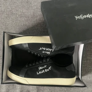 Svarta Saint Laurent sneakers strl 42 - Svarta sneakers från Saint Laurent med vit handskriven logga på sidan. Skorna har svart snörning, vit platt sula och detaljer i skinn och canvas. Klassisk och stilren modell med rund tå, perfekt för dig som gillar exklusiva streetwear-vibbar.