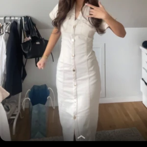 Vit långklänning med knappar - Snygg vit långklänning med korta ärmar och knappar hela vägen framtill. Klänningen har en figurnära passform och en liten slits nedtill. Perfekt för dig som gillar stilrena och eleganta plagg.