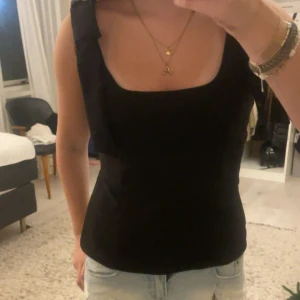 Svart linnetopp med rosett från Zara - Supersnygg svart linnetopp från Zara med breda axelband och en stor rosett på axeln. Toppen har en fyrkantig urringning och sitter figurnära. Perfekt att styla med jeans eller kjol för en trendig look.