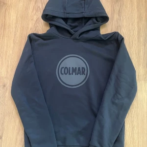 Svart Colmar hoodie med logga - Mörkblå hoodie från Colmar med stor tryckt logga på bröstet. Klassisk modell med huva och långa ärmar. Mjuk bomullskänsla och ribbade muddar. Perfekt för en chill och stilren look.          Tröjan är köpt på KidsBrandStore och är i storleken 164cm / 14 år. 