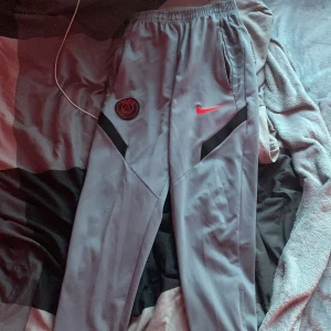 PSG x Nike grå träningsbyxor - Grå träningsbyxor från Nike i samarbete med Paris Saint-Germain. Byxorna har elastisk midja, svarta detaljer och PSG-logga samt röd Nike swoosh framtill. Perfekta för fotboll eller chill, med skön passform och sportig look. Storlek S