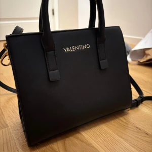 Svart handväska från Valentino - Stilren svart handväska från Valentino med guldiga detaljer och logga framtill. Väskan har både handtag och axelrem. Klassisk, fyrkantig form och minimalistisk design. Ordenarie pris 1200kr 