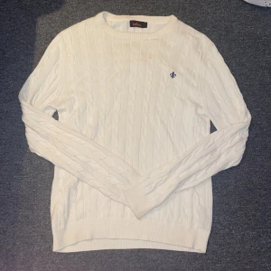 Vit kabelstickad tröja från Morris - Vit kabelstickad tröja från Morris med rund hals och diskret blå broderad logga på bröstet. Tröjan har lång ärm och ribbade muddar vid ärmslut och nederkant. Perfekt för dig som gillar klassisk och stilren look.