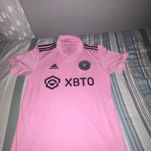 äkta Inter Miami Messi 10 Adidas tröja rosa - Säljer en officiell Inter Miami fotbollströja från Adidas i ljusrosa med svarta detaljer. Tröjan har korta ärmar, klubbmärke på bröstet, sponsortryck och Messi 10 på ryggen. Tillverkad i lätt och ventilerande material, perfekt för fotboll eller att bära som supporter. aldrig använd