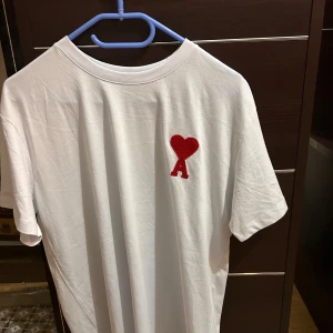 Vit t-shirt från AMI med rött hjärta - Vit t-shirt från AMI med klassisk passform och rund hals. På bröstet finns ett broderat rött hjärta med bokstaven A, vilket ger en snygg och unik detalj. Tillverkad i mjuk bomull som känns skön mot huden. Perfekt för en clean och stilren look.