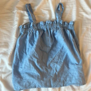 Ljusblå linnetopp med volangkant - Supersöt ljusblå linnetopp med breda axelband och volangdetalj upptill. Toppen har en avslappnad passform och är gjord i ett luftigt linnematerial, perfekt för varma dagar. Enkel och stilren design som är lätt att matcha med jeans eller kjol.