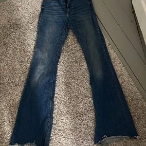 Blå bootcut jeans från Stradivarius - Snygga blå jeans från Stradivarius i bootcut-modell med utsvängda ben och klassisk femficksdesign. Jeansen har en mörkblå tvätt med lätt slitning och råa kanter nertill. Perfekta för dig som gillar retrovibbar och vill ha en cool siluett.