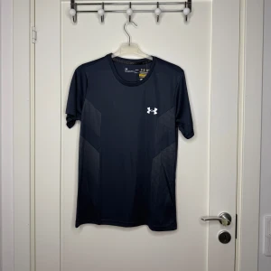 Mörkblå Under Armour träningst-shirt M - Snygg svart t-shirt från Under Armour med diskreta mönsterdetaljer och logga på bröstet. Tillverkad i polyester med HeatGear-teknologi som håller dig sval under träningen. Regular fit med korta ärmar och rund hals. Perfekt för gymmet eller löprundan.