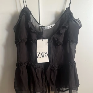 Svart volangblus från Zara - Säljer en svart, transparent blus från Zara med tunna axelband och flera volangdetaljer. Blusen har en luftig och flowy passform med rynkade sömmar som ger en romantisk känsla. Perfekt att styla med jeans eller kjol för en trendig look.
