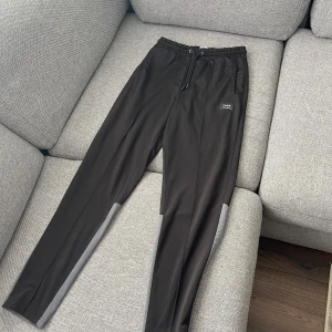 Svarta och grå mjukisbyxor från Solid - Svarta mjukisbyxor från Solid med grå paneler på nedre delen av benen. Byxorna har elastisk midja med snörning och sidofickor. Perfekta för chill eller träning, tillverkade i mjukt syntetmaterial för maximal komfort.