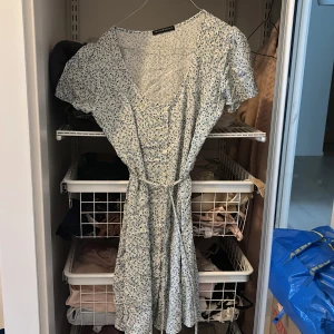 Blommig kortklänning Brandy Melville - Supersöt kortklänning från Brandy Melville i vit med små blå blommor. Klänningen har v-ringning, knappar framtill, korta ärmar och ett knytband i midjan. Man kan knyta den antingen på framsidan eller på baksidan för att göra den tajtare. Passar XS och S