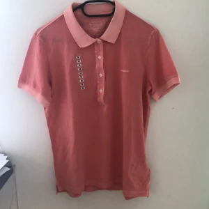 Peachy pink pikétröja från Gant - Snygg peachy pink pikétröja från Gant med klassisk krage och knappar framtill. Tröjan har korta ärmar och är tillverkad i bomull, perfekt för en avslappnad och sportig look. Gant-logga broderad på bröstet.