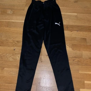 Svarta träningsbyxor från Puma S - Svarta träningsbyxor från Puma med elastisk midja och dragkedjefickor. Byxorna har en slim passform och är gjorda i ett glansigt syntetmaterial. Puma-logga i vitt på låret ger en sportig vibe. Perfekta för gymmet eller chill.