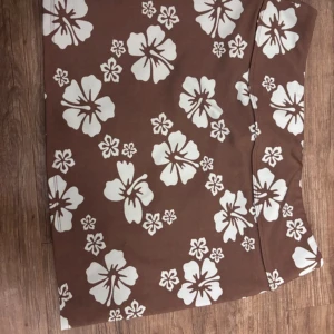 Brun blommig kortkjol - Snygg brun kortkjol med stora vita blommor i ett tropiskt mönster. Kjolen har en rak passform och är perfekt för dig som vill sticka ut med en somrig vibe. Materialet känns mjukt och bekvämt mot huden.