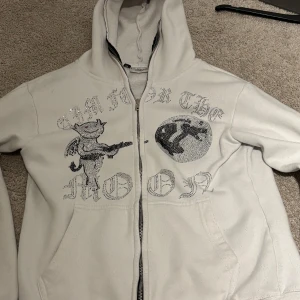 Vit hoodie Aim For The Moon  - Vit hoodie med dragkedja från Aim For The Moon. Framsidan har ett glittrigt tryck med text och en tecknad figur samt måne i strass. Tröjan har huva och är långärmad, tillverkad i mjukt material med fleece på insidan. Perfekt för dig som gillar streetwear med extra bling. Storlek M