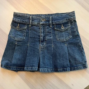 Plisserad jeanskjol med fickor - Snygg kort jeanskjol med plisserad nederdel och två fickor framtill med lock och knapp. Kjolen har bälteshällor och klassisk jeanslook i mörkblå denim. Perfekt för en cool och avslappnad stil. Populär så var snabba!!