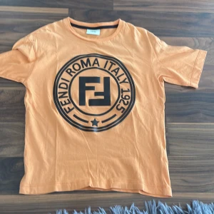 Orange Fendi t-shirt med stor logga - Snygg orange t-shirt från Fendi med stor svart logga och texten 'Fendi Roma Italy 1925' på bröstet. Klassisk rund hals och korta ärmar. Tillverkad i mjuk bomull som är skön att bära. Perfekt för dig som gillar exklusiva streetwear-vibbar.Från NK i Stockholm