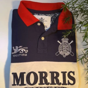 Morris Cricket pikétröja marinblå röd - Snygg pikétröja från Morris med marinblå bas, röd krage och vit panel med broderad text 'Morris Cricket'. Detaljer som klubbmärke, lejon och siffran 68 på ärmen. Klassisk krage och knappar framtill. Perfekt för dig som gillar sportig och preppy stil.