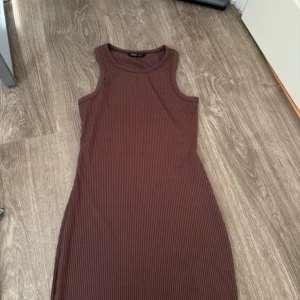 Ribbad brun ärmlös klänning - Säljer en ribbad brun klänning från Lindex. Modellen är ärmlös med rund halsringning och har en tight passform som framhäver figuren. Perfekt för dig som gillar en enkel men stilren look.