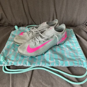 Nike Mercurial ZM Vapor 16 elite FG  - Snygga Nike Mercurial fotbollsskor i silver med neonrosa detaljer och logga. Skorna har snörning, låg profil och är tillverkade i syntetmaterial för bästa känsla på planen. Sulan har turkosa dobbar och cool design. Perfekta för dig som vill sticka ut på fotbollsplanen.