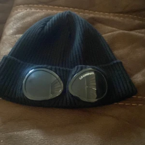 Svart goggle-mössa från C.P. Company - Svart ribbstickad mössa från C.P. Company med ikoniska goggle-linser framtill. Mössan är tillverkad i mjukt material och har en tight passform. Perfekt för dig som vill sticka ut med streetwear-vibe och unik design.