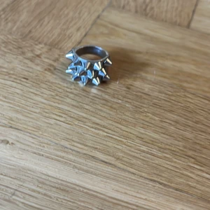 Silvrig ring med nitar - Cool och chunky ring i silverfärgad metall med flera spetsiga nitar runt hela bandet. Perfekt statement piece för dig som gillar edgy och alternativ stil. Ringen har en bred form och ger en riktigt tuff vibe.