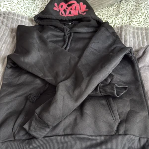 Syna world - Svart hoodie och byxor med stor huva och snörning, prydd med ett fett röd graffiti-tryck på huvan. Tröjan har ribbade muddar och känguruficka framtill. Perfekt för dig som gillar streetstyle och vill sticka ut med coola detaljer